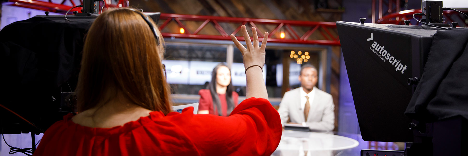 A student cues two television news anchors from behind the camera.